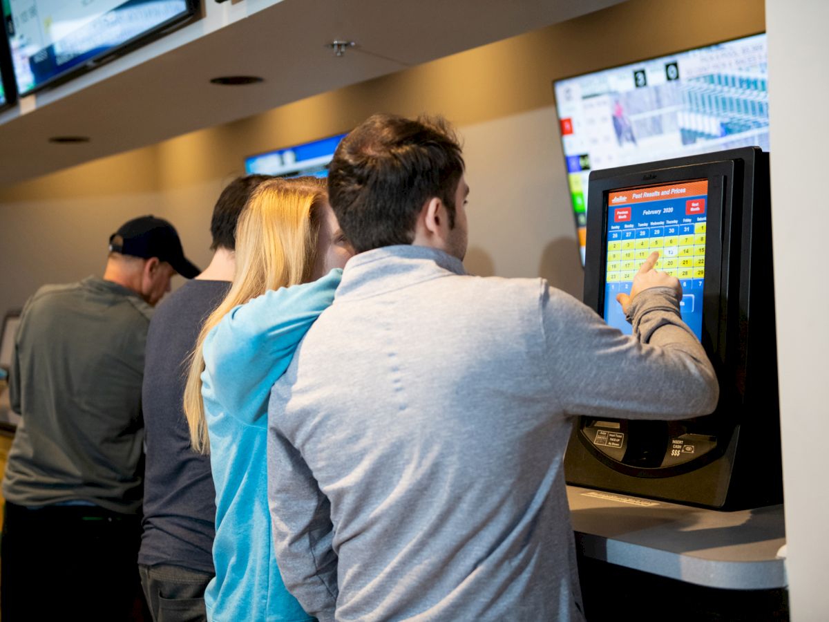 People using self-service betting terminals, browsing and placing bets, with multiple screens displaying sports events in the background.