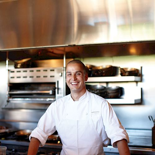 A chef is standing in a professional kitchen, smiling, and leaning on a counter. He is wearing a white chef's uniform and apron.
