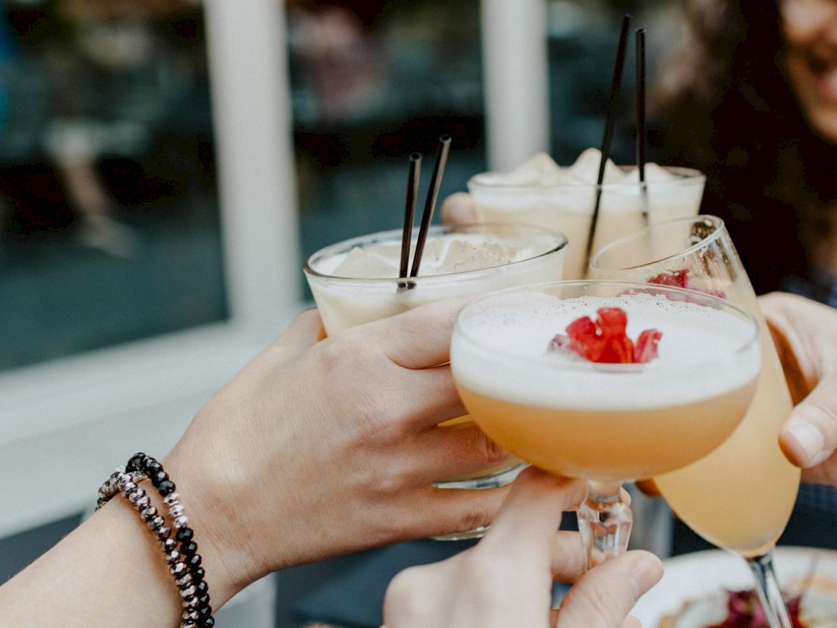 Several people are clinking their cocktails together in a celebratory toast, with visible fruit garnishes and straws in the drinks.