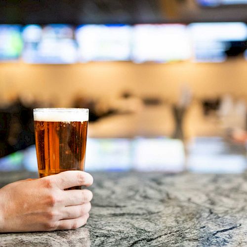 A hand is holding a glass of beer on a granite counter in a bar, with blurry screens and ambient lighting in the background.