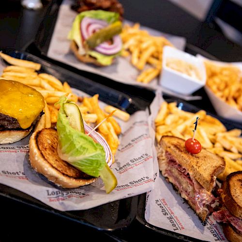 The image shows trays with burgers, fries, and a sandwich with a side of fries on a black surface.