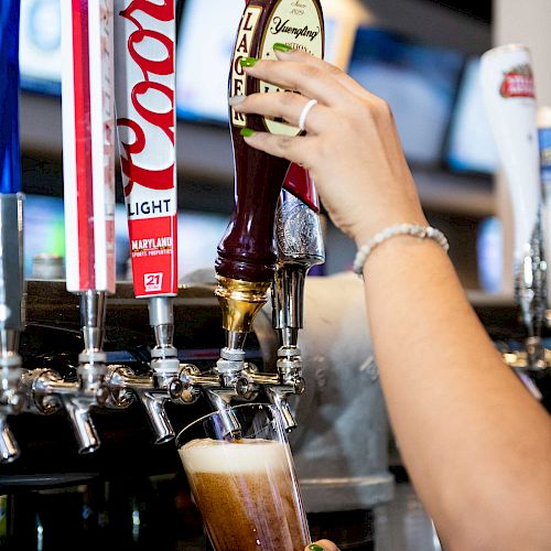 A person's hand pours beer from a tap into a glass at a bar, with various beer tap handles visible, including Coors Light.