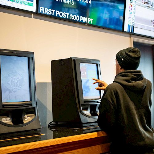 A person in a dark hoodie and beanie is using a touchscreen terminal, possibly for betting, with screens displaying race information above.