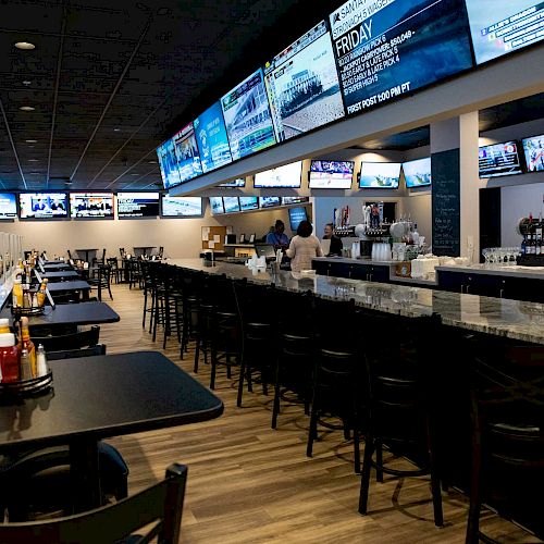 A spacious sports bar with numerous TV screens, a long bar counter, tables with condiments, and a person in the background near the bar.