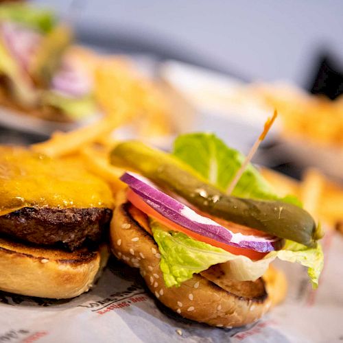 An open burger with cheddar cheese, lettuce, tomato, red onion, and a pickle, surrounded by French fries on a table.