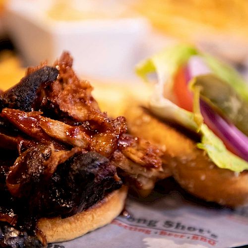 The image shows a delicious sandwich topped with pulled pork and accompanied by vegetables like lettuce, tomato, and onions on the side of the bun.