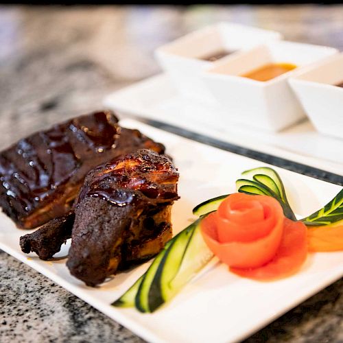 The image shows a plate with glazed meat, garnished with a vegetable rose and cucumber slices, accompanied by dipping sauces in small bowls.