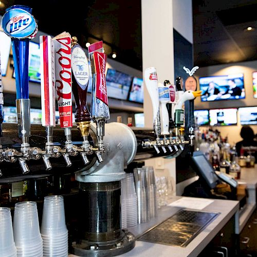 This image shows a bar with multiple beer taps, plastic cups, and a series of TV screens in the background.