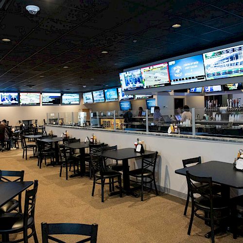 The image shows a sports bar with multiple TV screens, black tables and chairs, and a counter with taps. Condiments are on the tables.