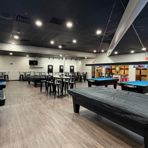 The image shows a billiards hall with several pool tables, a dart area, and seating, with a modern interior and overhead lights.