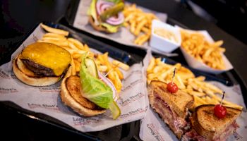 The image shows trays with cheeseburgers, sandwiches, and fries, each tray lined with paper. There is also a small square bowl with a dip or sauce.