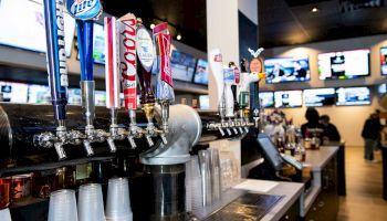 A bar with multiple beer taps, plastic cups, and TV screens showing sports in the background. Some people are seen near the counter.