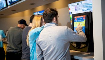 A group of people is using touchscreen kiosks, possibly for betting or gaming, inside a modern establishment with screens displaying various information.