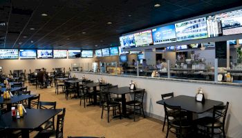 The image shows a sports bar with multiple TV screens displaying various channels, tables and chairs arranged for dining, and a bar area in the background.