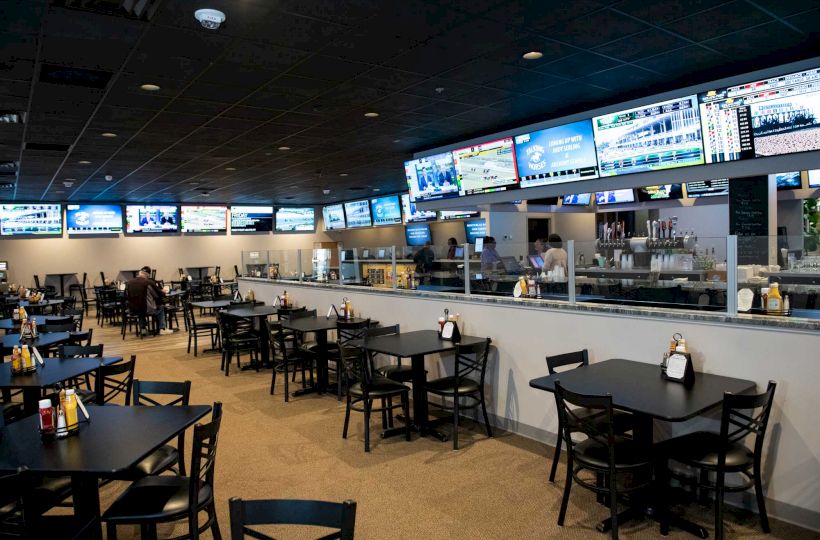 The image shows a sports bar with multiple TV screens displaying various channels, tables and chairs arranged for dining, and a bar area in the background.