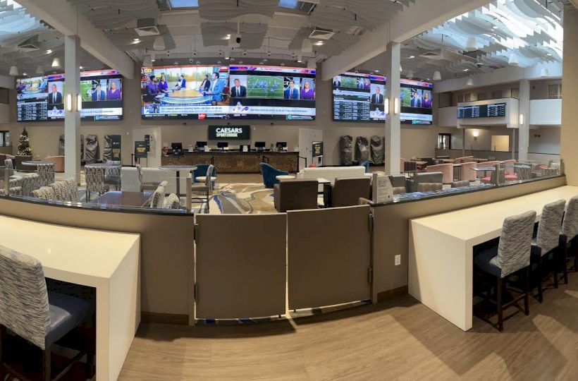 The image shows a spacious, modern sports betting area with multiple TV screens, seating, and an open, airy design.