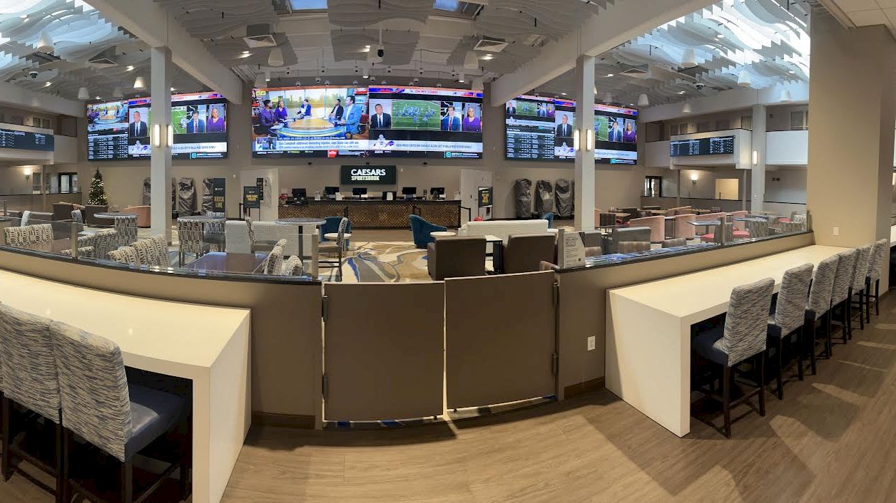 The image shows a spacious, modern sports lounge with multiple large screens displaying sports channels and many seating areas.