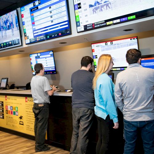 People are at kiosks in a betting area, with multiple screens displaying odds and events above them.