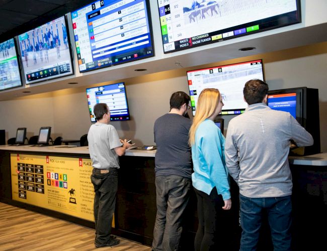 People are at kiosks in a betting area, with multiple screens displaying odds and events above them.