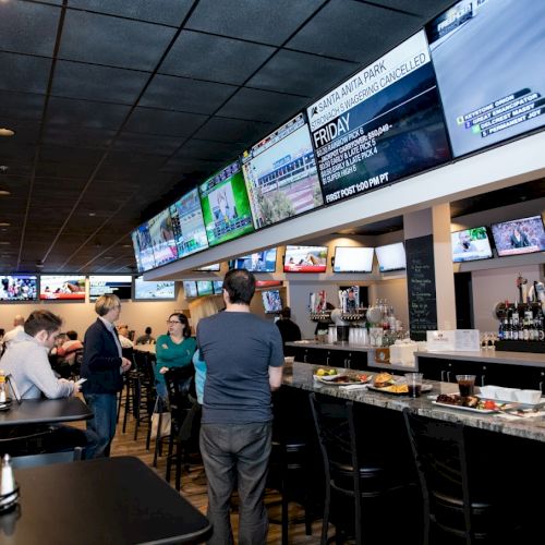 A sports bar with multiple large screens showing various games, people at the bar, drinks, and food on tables.