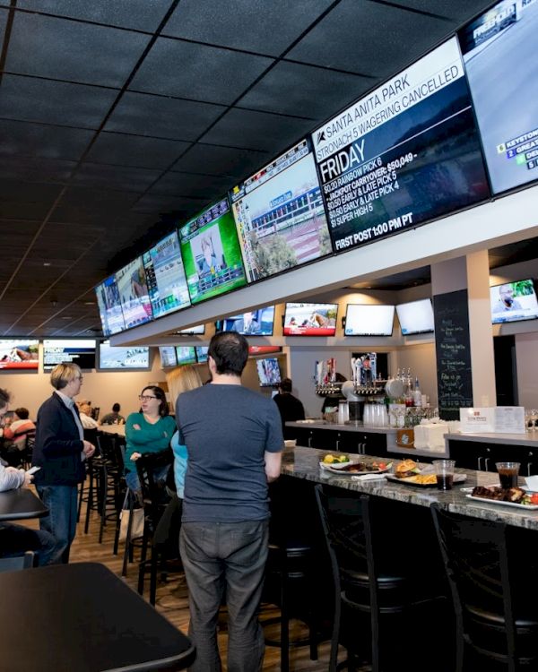 A sports bar with multiple large screens showing various games, people at the bar, drinks, and food on tables.