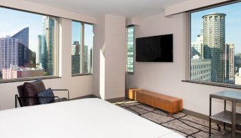 This image shows a modern hotel room with large windows, a bed, and a wall-mounted TV overlooking a cityscape.