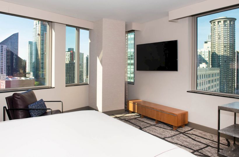 This image shows a modern hotel room with large windows, a bed, and a wall-mounted TV overlooking a cityscape.