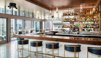 A modern, stylish bar with high shelves stocked with a variety of bottles, blue cushioned stools, and large windows offering a city view.