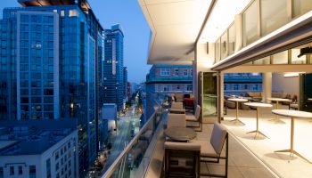 A modern balcony with seating overlooks a city street and tall buildings during the evening, with interior furnishings and lighting visible.