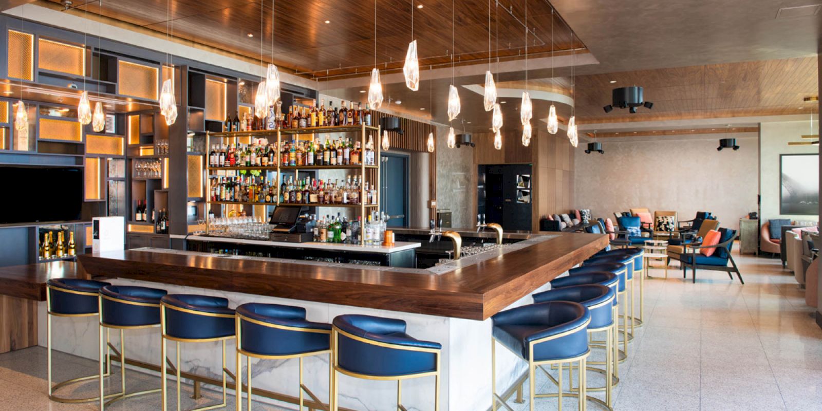 The image shows a stylish bar with a wooden counter, blue bar stools, hanging lights, and a well-stocked shelf of bottles.