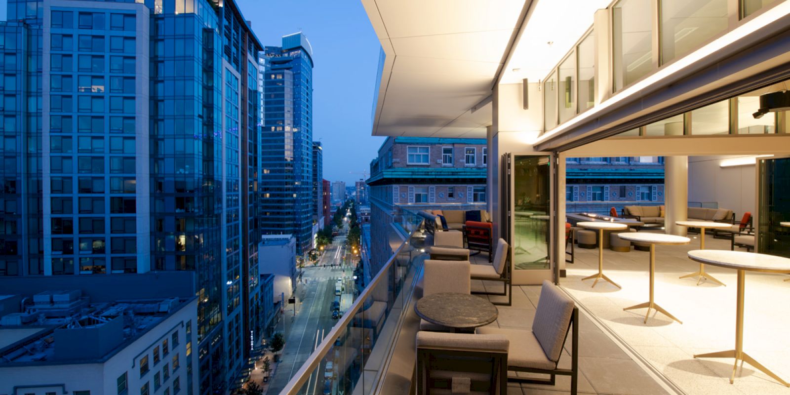 A modern balcony overlooking a cityscape at dusk, featuring outdoor seating with tables and chairs, and buildings illuminated by street lights.