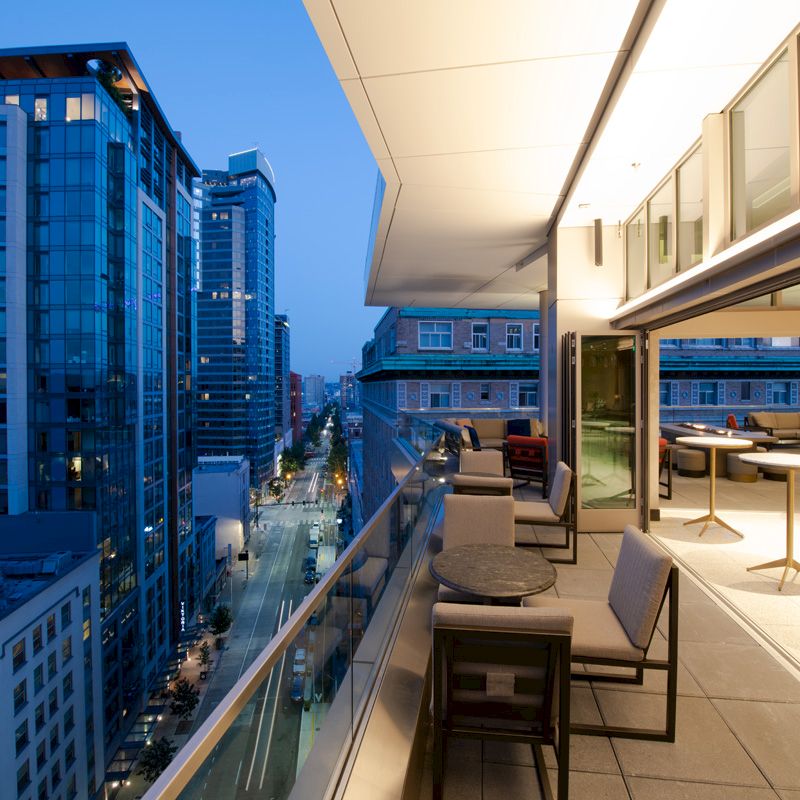 The image shows a modern cityscape during dusk, featuring tall buildings and a balcony with outdoor furniture and tables viewed from a high vantage point.