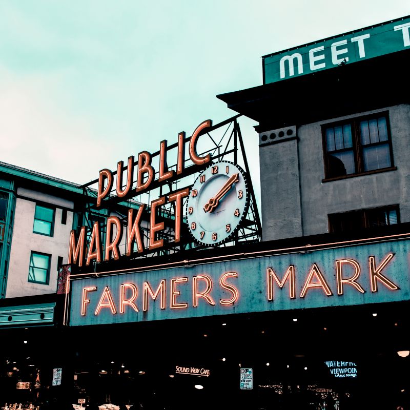 Image of a farmers market with prominent neon signs displaying "PUBLIC MARKET" and "FARMERS MARKET" in front of urban buildings.