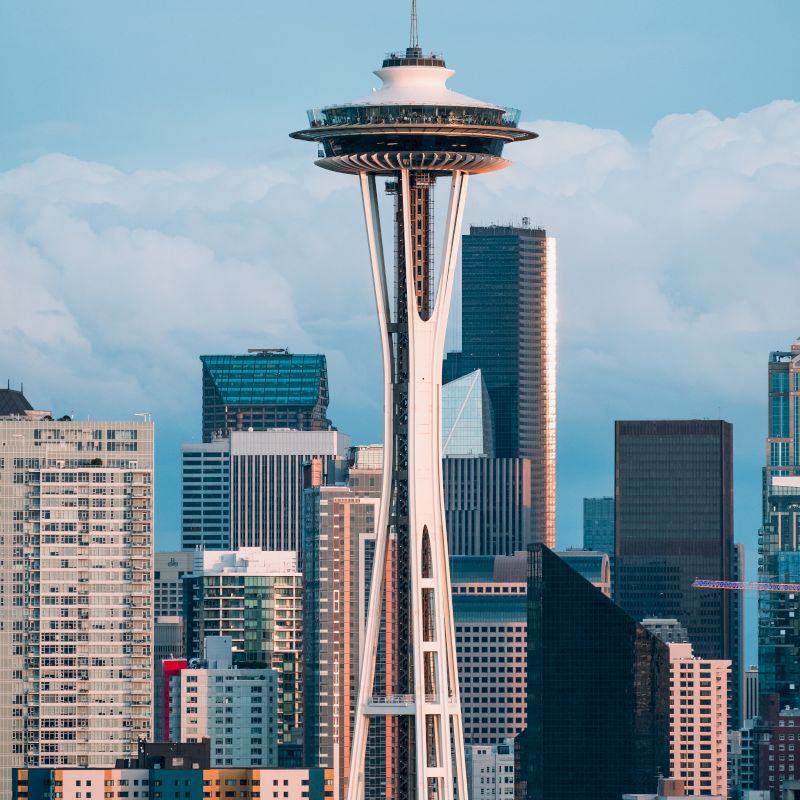 The image shows the iconic Space Needle against a background of modern skyscrapers and buildings, under a partly cloudy sky.