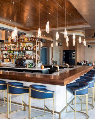 A modern bar with blue barstools, a well-stocked liquor shelf, and hanging pendant lights. Lounge seating is visible in the background.
