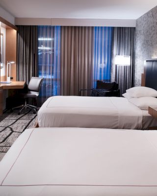 A modern hotel room with two neatly made beds, a TV on the wall, a desk and chair near the window, and stylish lighting and decor.