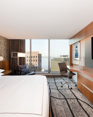 This is a modern hotel room featuring a large bed, desk, chair, wall-mounted TV, and a view of buildings through a window.