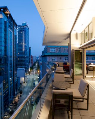A modern cityscape at dusk with tall buildings and an open balcony featuring seating and tables, overlooking a bustling urban street.
