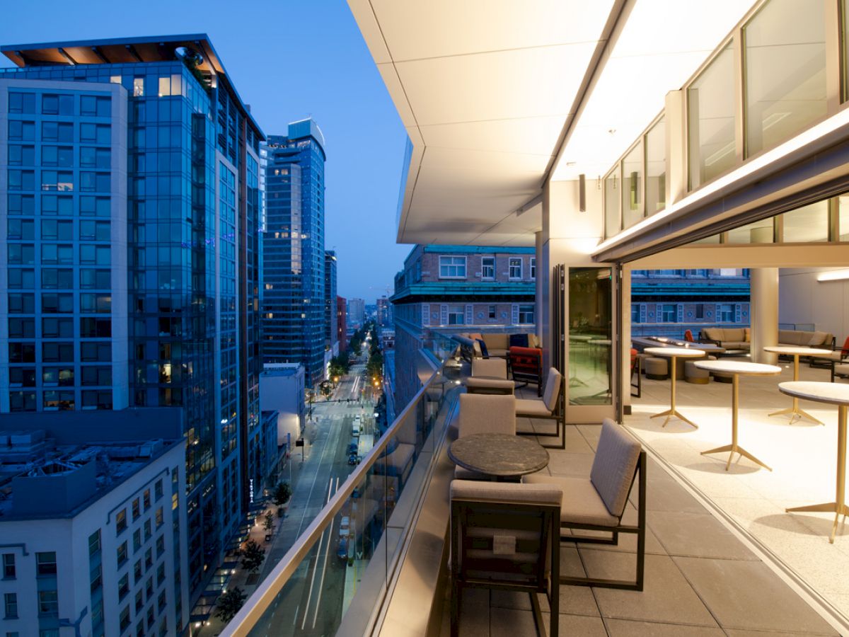 A modern balcony with seating overlooks a cityscape with tall buildings. The scene is during the evening with the balcony and city lights on.