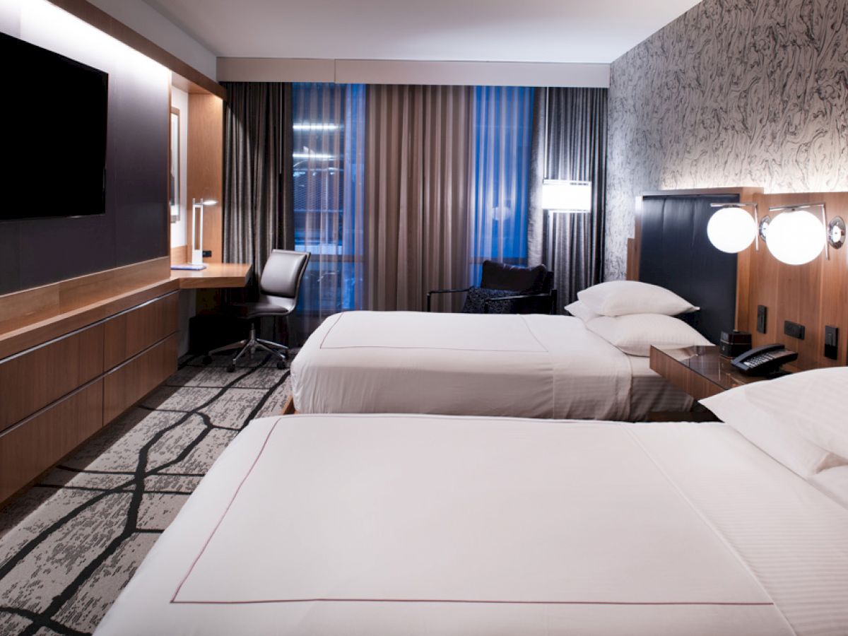 The image shows a modern hotel room with two neatly made beds, a wall-mounted TV, a desk with a chair, and stylish lighting and decor.