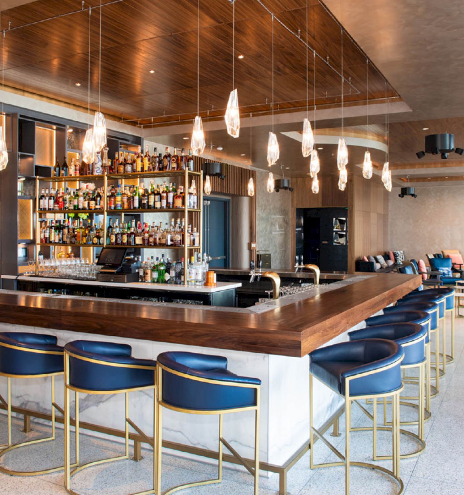 A modern bar with a wooden counter, blue bar stools, hanging lights, and a fully stocked liquor shelf in a stylish, spacious setting.