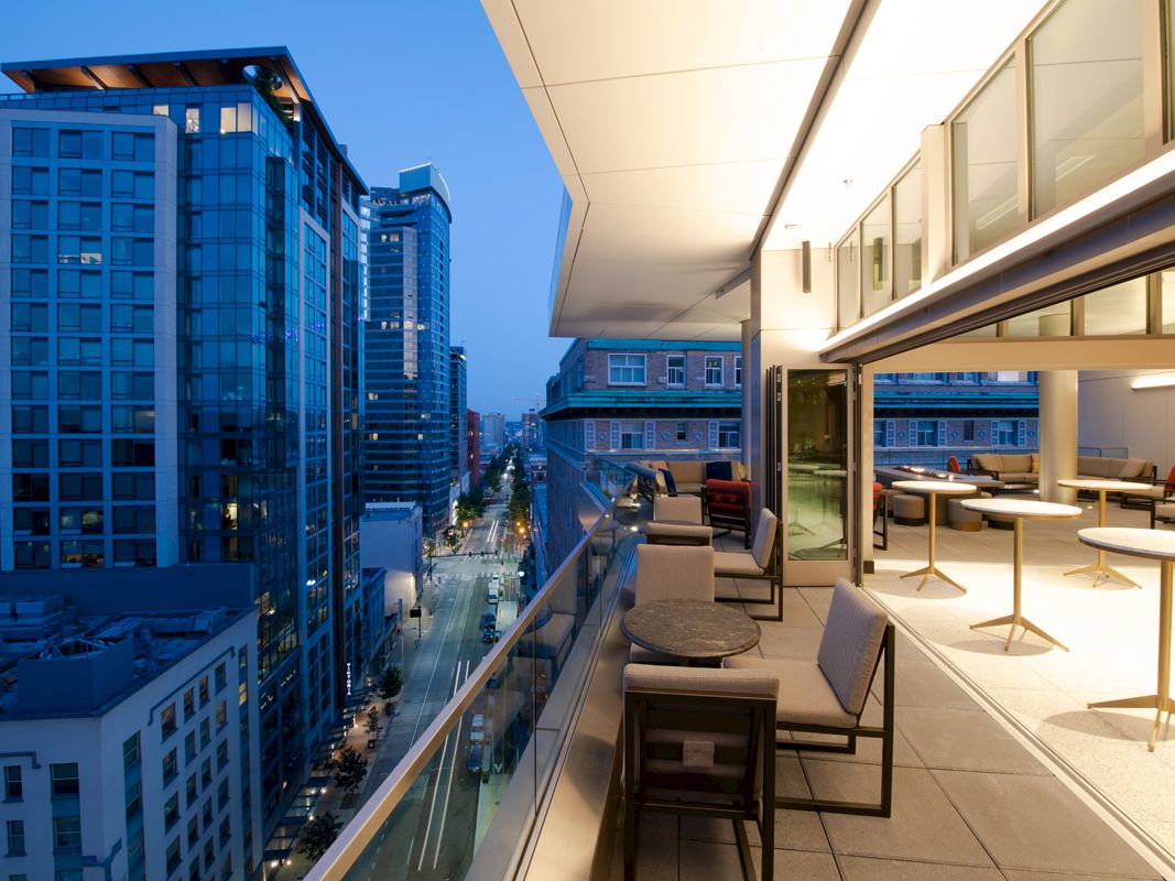 A modern cityscape view from a high-rise balcony at dusk, featuring seating areas and tall buildings along a vibrant street below.