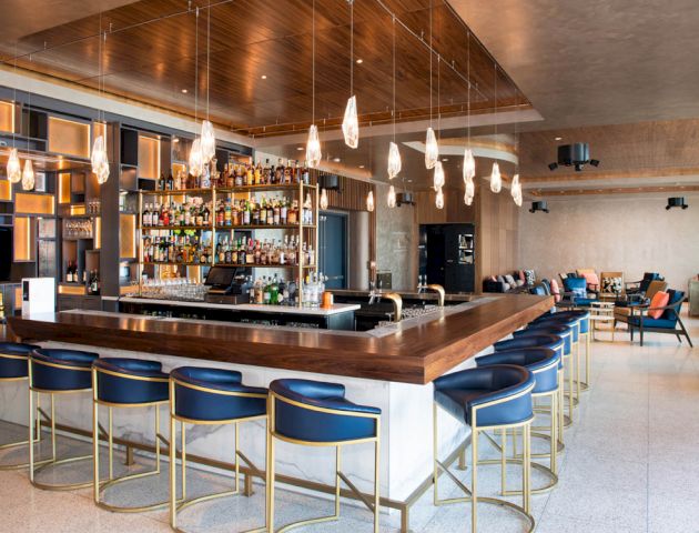 The image shows a modern bar with blue bar stools, a well-stocked back bar, hanging lights, and a seating area with chairs and tables.