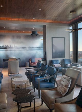 A modern lounge with ample seating, large windows offering a scenic waterfront view, and a minimalist design featuring a mural of a city skyline on one wall.