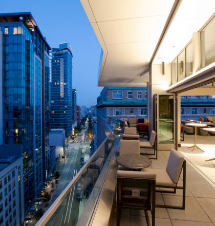 The image shows a modern cityscape with tall buildings viewed from a balcony at dusk, featuring outdoor seating and a glass railing along the edge.