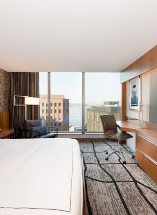 A modern hotel room with a large bed, a work desk, a TV on the wall, and a window with a city view.