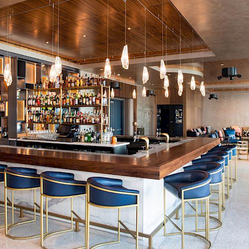 This image depicts a modern bar with a wooden counter, blue stools, hanging light fixtures, a stocked bar shelf, and seating areas in the background.
