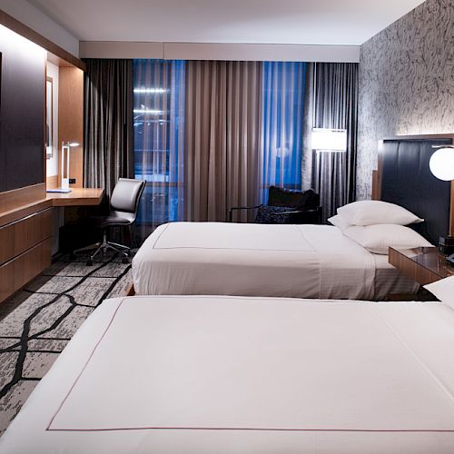 A modern hotel room with two neatly made single beds, a wall-mounted TV, a long desk with a chair, and stylish lighting.