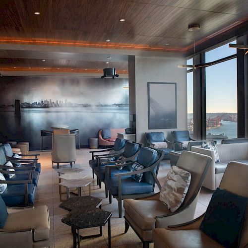 The image shows a modern lounge with multiple chairs and tables, large windows offering a scenic view of water and a Ferris wheel in the background.