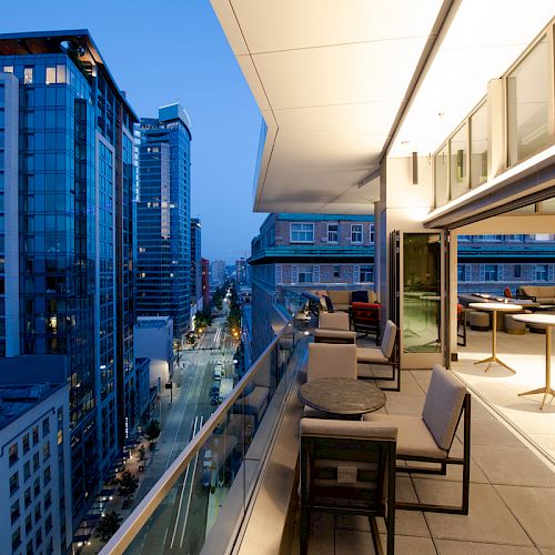 A modern city view with tall buildings during dusk is seen from an open balcony with seating and tables. The balcony overlooks the street below.